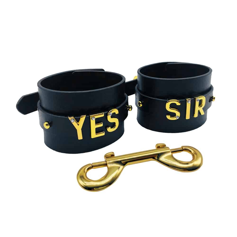 Pair of handcuffs for wrists personalized with writing YES SIR on black Italian leather with letters and hangers in 24 carat gold plated and small stones on each of the letters from the collaboration UPKO X Brigade Mondaine presented on a white background at Brigade Mondaine