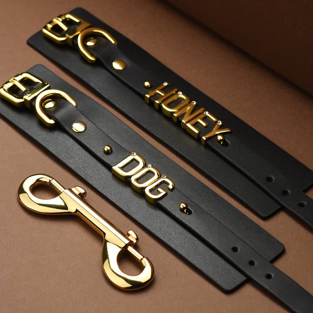 Pair of soft black Italian leather handcuffs laid flat with the words DOG and HONEY in 24 karat gold plated letters with stone inlaid on each letter all laid flat on a brown background with a light coming from the top left corner of the image being ideas for the UPKO X Brigade Mondaine collaboration handcuffs