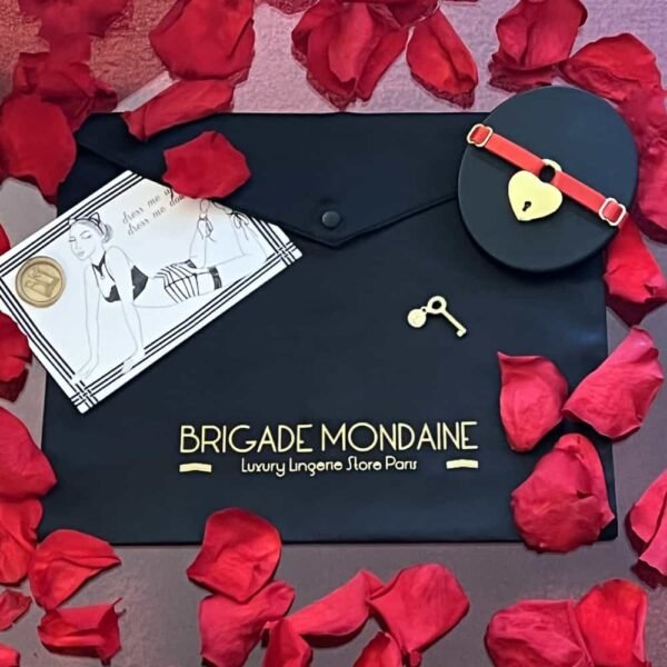 black packaging of the brigade mondaine. On the fabric is inscribed the brigade mondaine of golden color. Accompanied by a card, a red choker and rose petals.