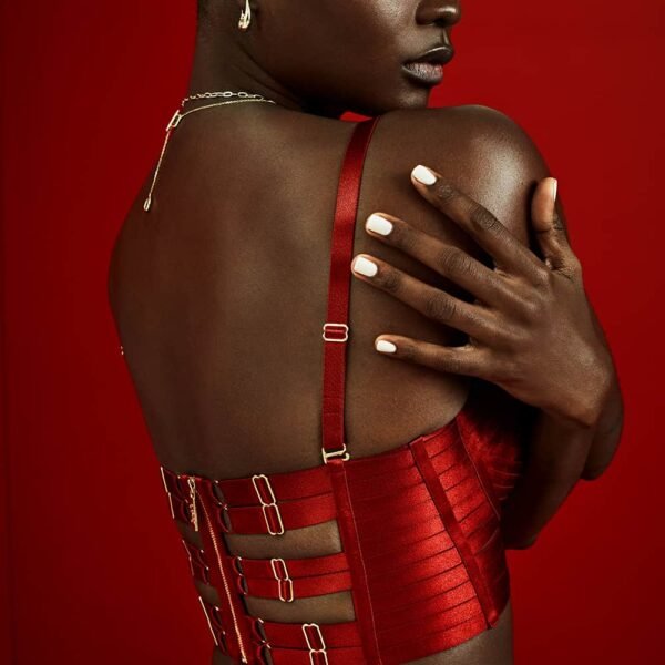 The model is wearing a Signature Bordelle set in red. It is composed of a Minerva Ruby bustier and an Exclusive Classic Waspie Ruby, both made of red satin and fishnet elastic and gold plating.