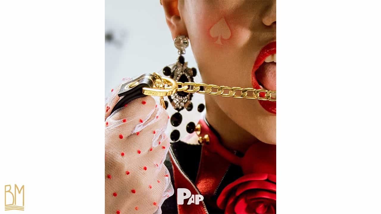 PAP Magazine it is a woman carrying in her hand the leash of the brand Upko and around her neck she wears the Gag Ball Upko. She has white gloves with red dots and red lipstick. On her cheek, there is the sign of the spade.