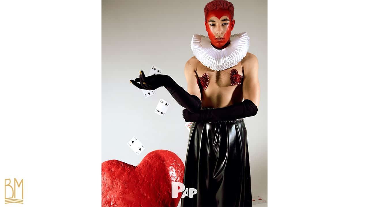 PAP Magazine it is a man throwing cards. His face is painted in red and forms the sign of the heart. He wears black gloves up to his forearms and Nippies of the brand Ruth Melbourne. Behind him there is a big red heart.