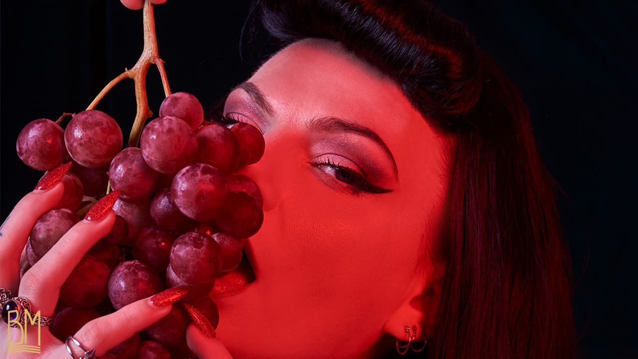 Foto de Julie Von Trash por Studio Volua Paris. Sostiene un gran racimo de uvas rojas en su mano derecha y mira al objetivo. La foto sólo muestra su cara y en particular acentúa su mirada.