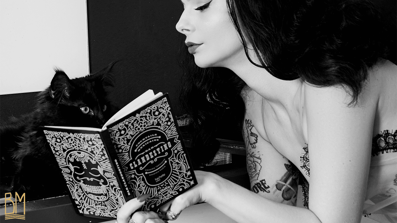 Foto de Julie Von Trash en blanco y negro por Studio Volua Paris. Lleva un kimono de la marca Ludique Lingerie. Está tumbada y leyendo un libro. A su derecha, hay un gato negro.